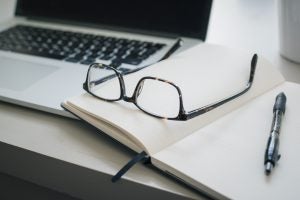 a pair of glasses and a pen on top of an empty notebook, all lying on a table near a laptop