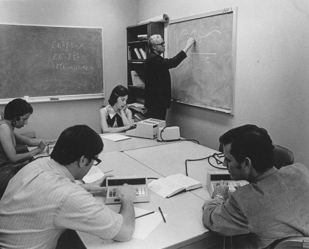 professor writing on a chalkboard, while students work on a calculator